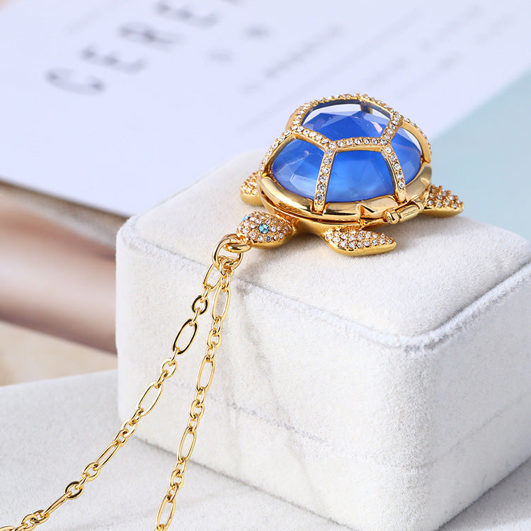 A blue and gold turtle pendant sitting on a white jewelry box